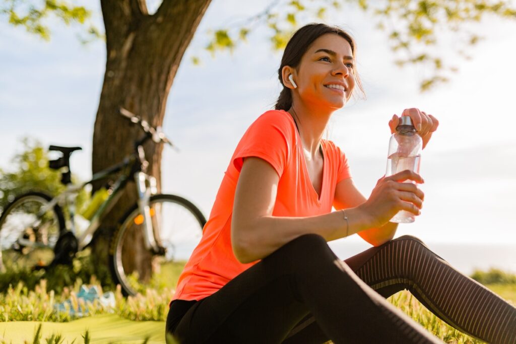 smiling beautiful woman drinking water in bottle doing sports in morning in park nature doing yoga on mat, pink fitness outfit, happy healthy lifestyle, music in earphones, bicycle on background